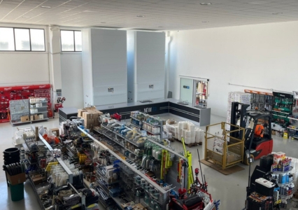 A bright warehouse is equipped with several Modula Lift vertical storage units, one showing accessible inventory and a worker at its controls.