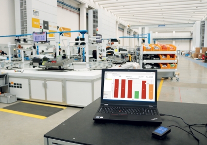 A laptop displays a progress bar chart on a black table in the foreground, overlooking a spacious, bright Modula warehouse.