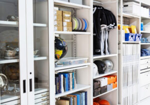 A white 4-Post Freespan™ Shelving system with many adjustable metal shelf levels filled with numerous types of merchandise including hanging shirts, stacked shoeboxes, basketballs, helmets, and books. A partial section of the system on the left is a glass-enclosed display case with a series of lower drawers.