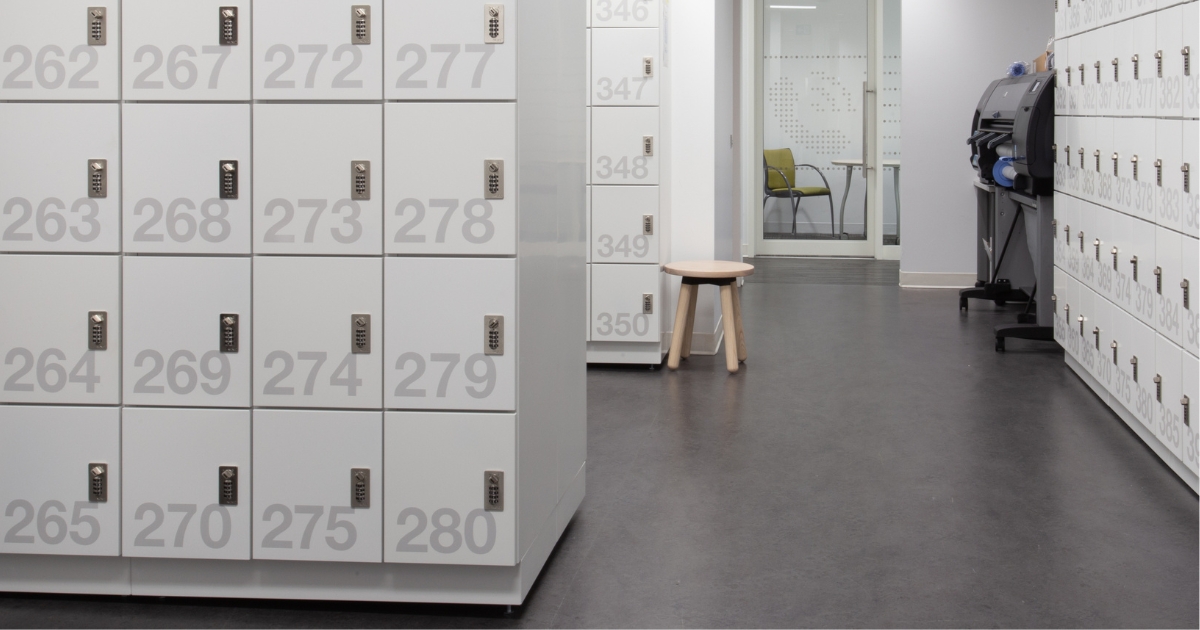A set of light grey office day use lockers by SpaceSaver with large gray numbers and small digital locks. The image focuses on the lockers in the foreground, with more lockers and a glass-walled room visible in the background.