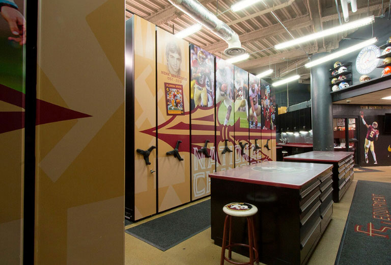 Education-Featured-Image University football equipment room with a mobile storage system decorated with team graphics. Dark work tables are in the foreground, and shelves displaying helmets line the back wall.