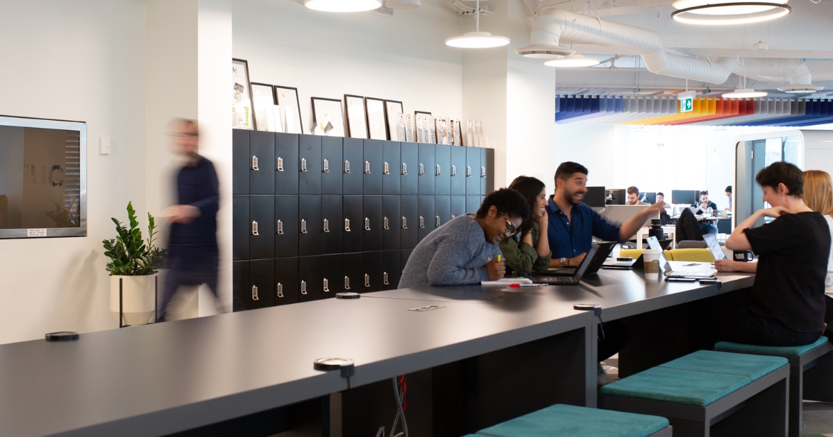 An open-concept office with a long communal desk where multiple people are working and collaborating. A wall of black day use lockers by SpaceSaver stands in the background under framed art.