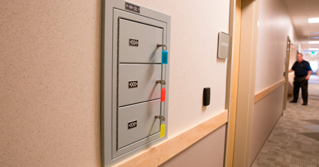 Gun lockers are mounted on a wall in a police station hallway. Each light grey locker has a number and a key with a coloured fob hanging from its lock.