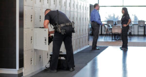 A police officer puts an item into one of many light grey personal storage lockers with mechanical locks, arranged along a wall. Two other officers stand nearby, conversing.