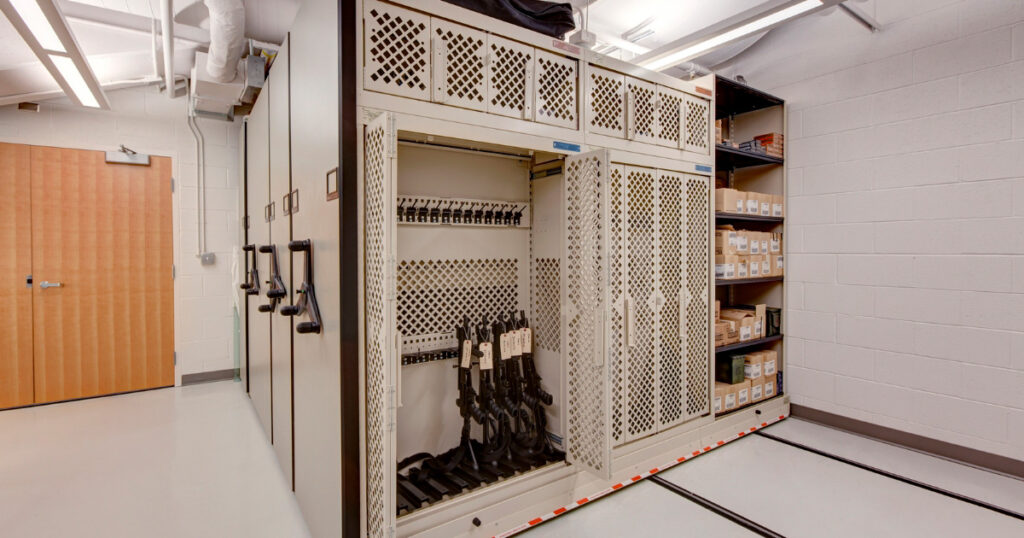 A beige weapons rack is incorporated into a mobile storage system, featuring shelves and compartments with various items. Several rifles are stored in the open section of the weapons rack.