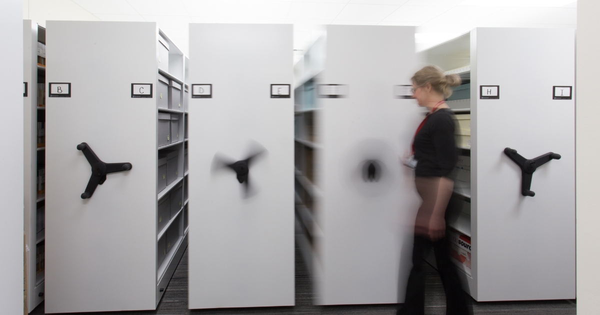 A motion-blurred shot of a woman walking past a row of light grey SpaceSaver mobile storage units in an office. The units, which are used for filing and storage, have letters on the front (B, C, D, F, H, I) and are equipped with large black handles for moving them along tracks.