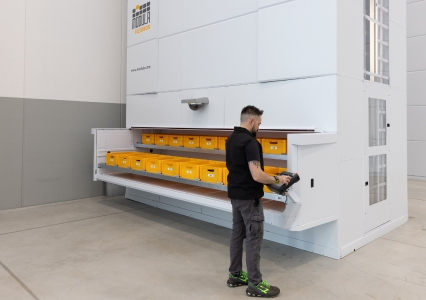 An eye-level view of the Modula Flexibox vertical lift module featuring a picking area with 9 bins per tray. A worker is standing on the right end of the picking bay and manages the system through the integrated copilot screen.