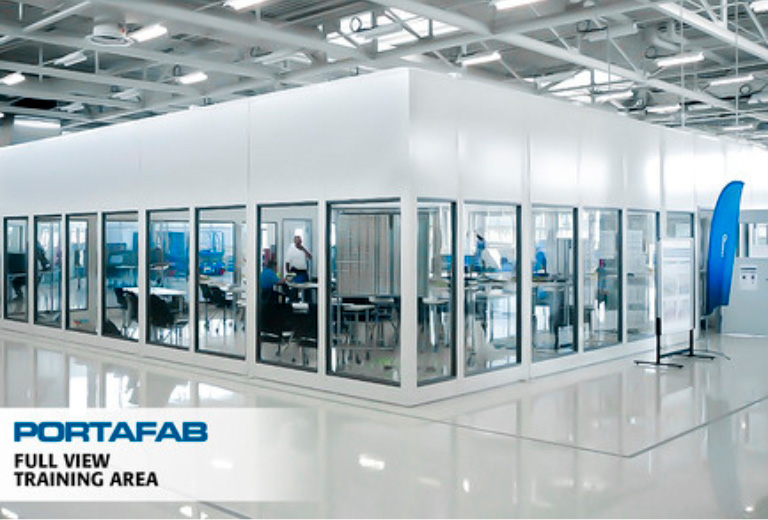 A brightly lit, modular building with glass walls encloses a training area within a larger industrial space. A text overlay reads ‘PORTAFAB FULL VIEW TRAINING AREA’.