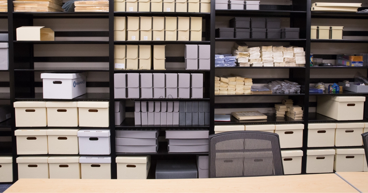 A wall of black 4-post shelving is filled with various sizes and colours of archival boxes and files. A desk and office chair are visible in the foreground.
