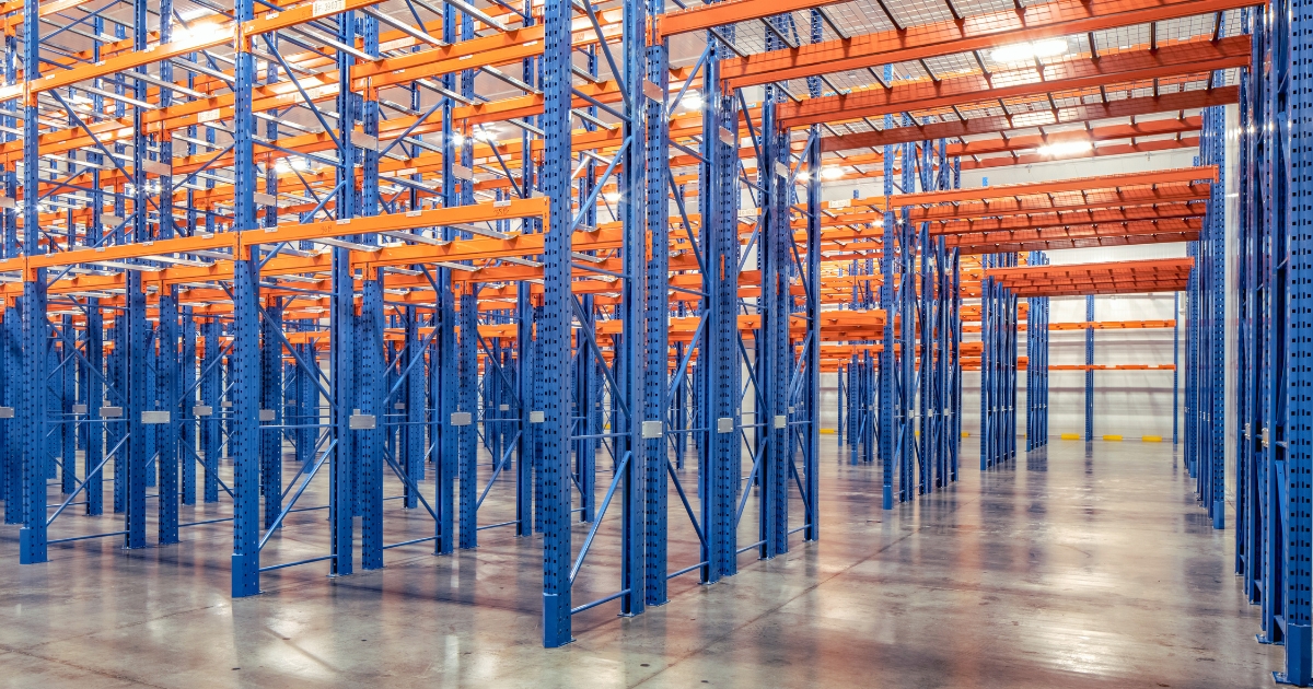 Racking-Shelving-Parts-Guide-Featured-Image Blue and orange pallet racking system with visible vertical uprights, horizontal load beams, safety bars, and overhead wire mesh deck parts in an empty warehouse.