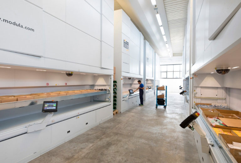Bright warehouse aisle lined with multiple tall, white Modula Vertical Lift Modules (VLMs). A worker operates one unit, while a tray of cardboard boxes is presented in the foreground retrieval bay.