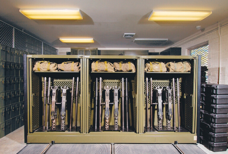 Public-Safety-Featured-Image Secure, olive green Universal Weapons Racks (UWRs) mounted on a high-density mobile storage system in an armory, storing multiple rifles and tactical gear bags.