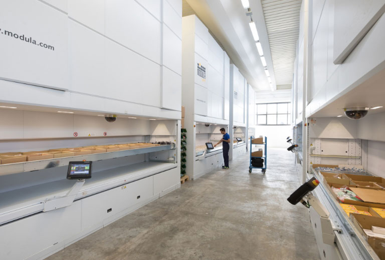 A bright warehouse is equipped with several Modula Lift vertical storage units, one showing accessible inventory and a worker at its controls.