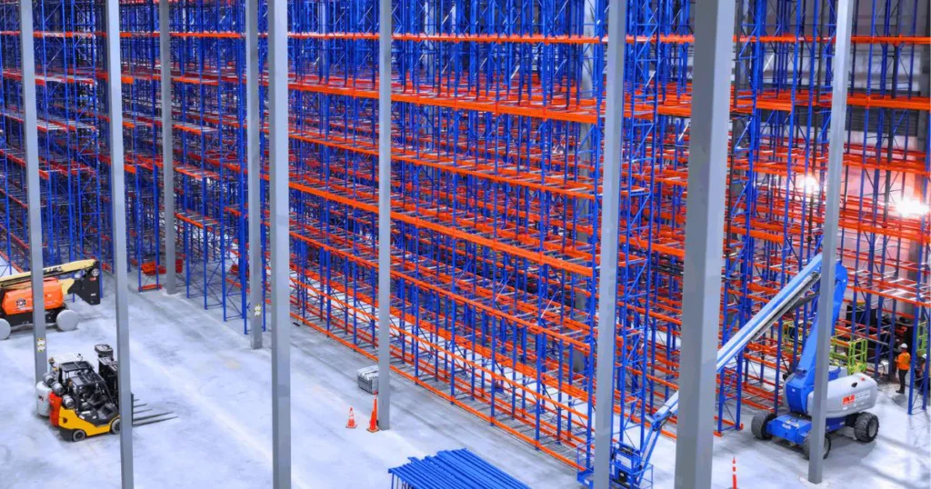 Massive warehouse interior during a pallet racking installation, featuring towering rows of blue and orange industrial steel racking with a forklift on the concrete floor.